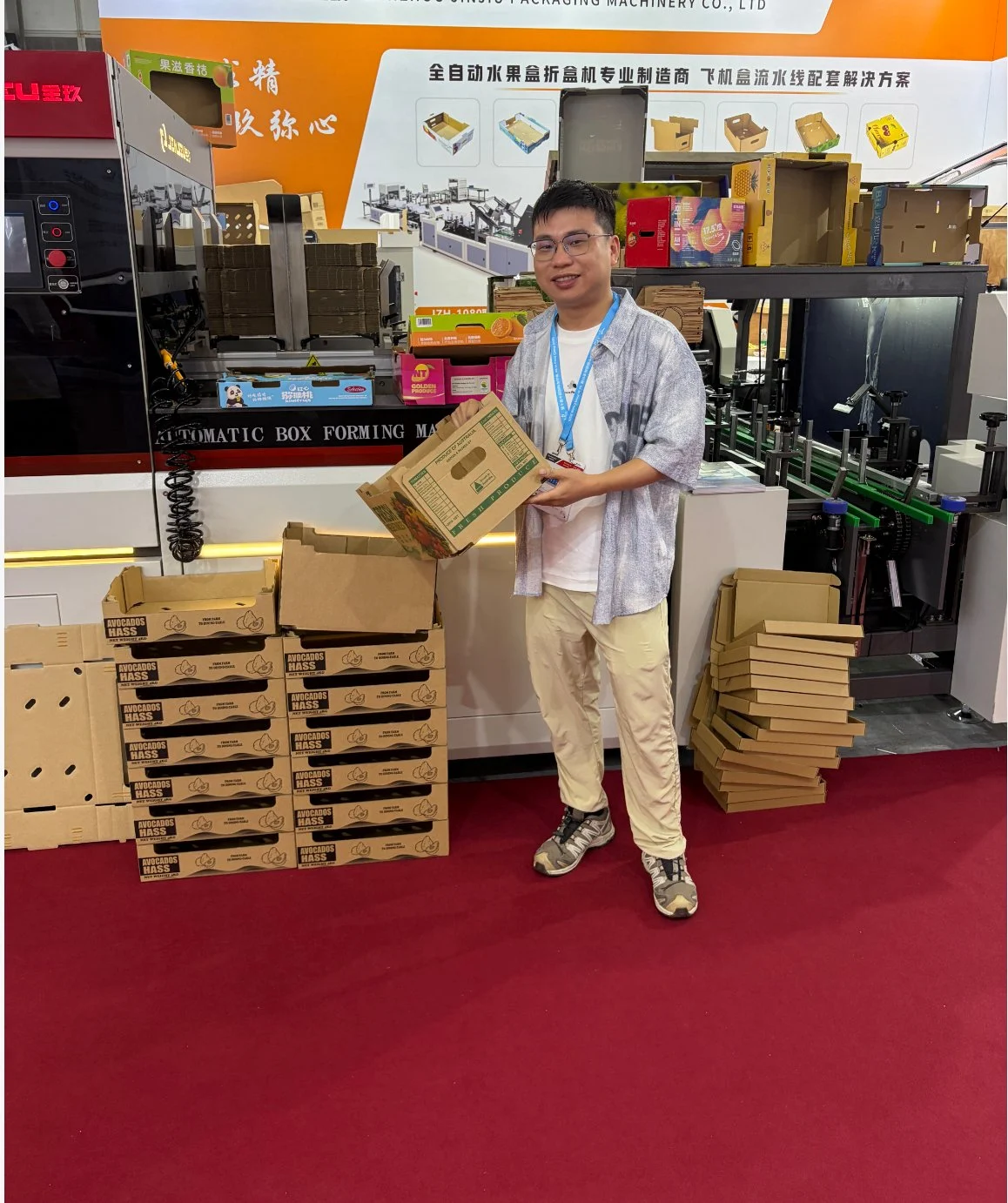 Una persona con in mano una scatola di frutta stampata accanto a una pila di campioni di scatole di avocado presso lo stand Xunding nel report XD-Z07 All in Print China 2026