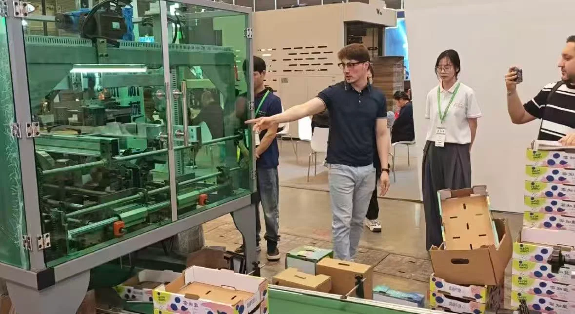Visitors looking at a guarded box forming machine and printed fruit trays at an exhibition