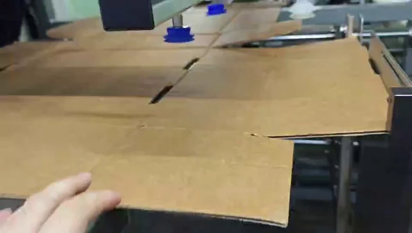 Flat die-cut cardboard blank positioned below the machine pickup area during the box-type changeover demonstration