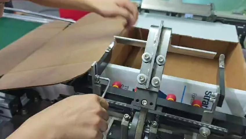 Technician adjusting the lower forming fixture beside a brown cardboard blank with an Allen key