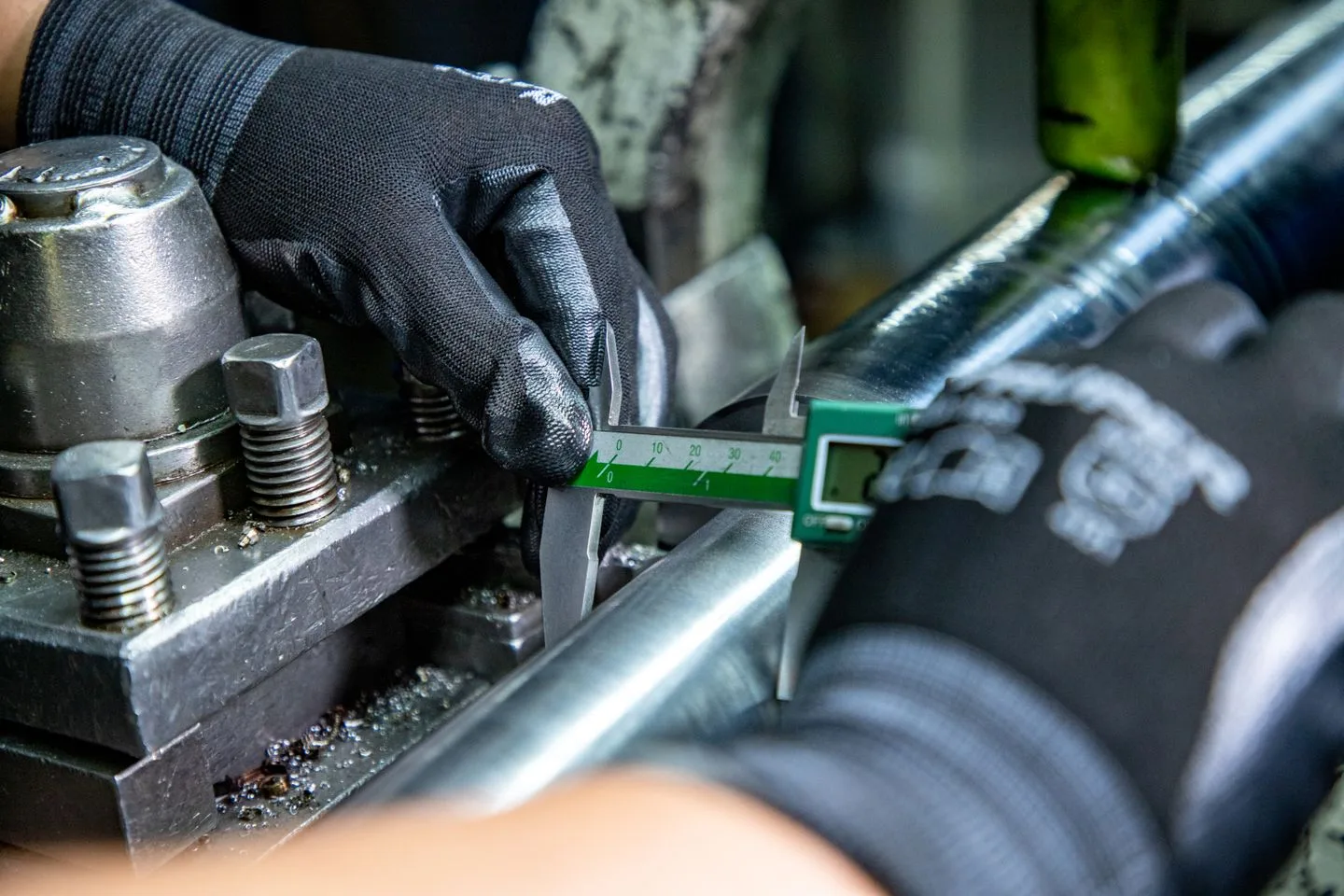 Gloved hands using the outside jaws of a digital caliper to measure a metal shaft on a stationary lathe